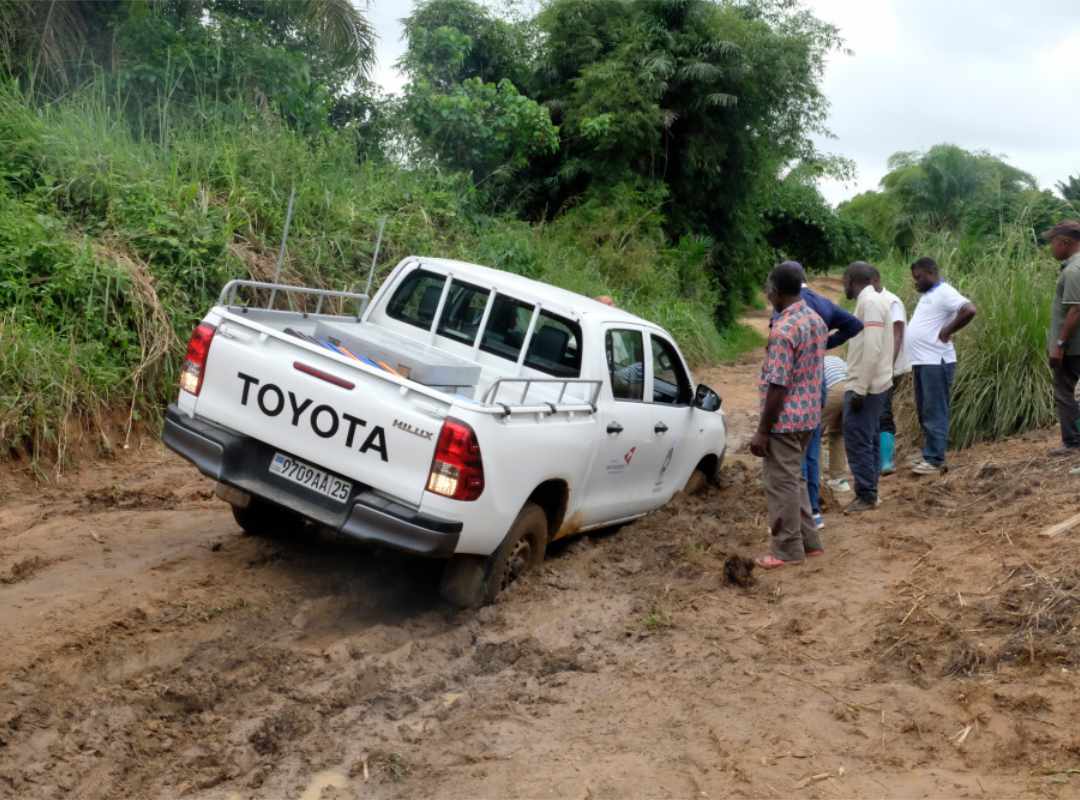 De auto van Kisangani developpement asbl is vastgeren in de modder na een fikse regenbui. verschillende mensen kijken toe.